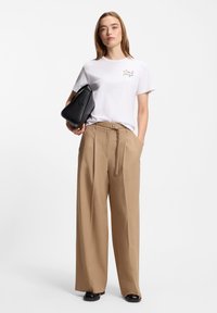 Άσπρη μπλούζα με κεντήματα λουλουδιών και μπεζ παντελόνι με φαρδιά πόδια. Το μοντέλο κρατάει μαύρη τσάντα. Λείο ύφασμα, άνετη εφαρμογή, ουδέτερη παλέτα χρωμάτων.