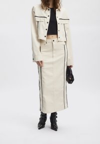 Beżowa kurtka dżinsowa z czarnym obszyciem, krótki fason; dopasowana długa spódnica z bocznymi paskami. Modelka nosi czarne botki na obcasie i małą torebkę.