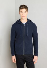 Felpa con zip di colore navy in un tessuto a maglia testurizzato, maniche raglan, cappuccio con cordino e zip liscia; indossata sopra una maglietta azzurra.