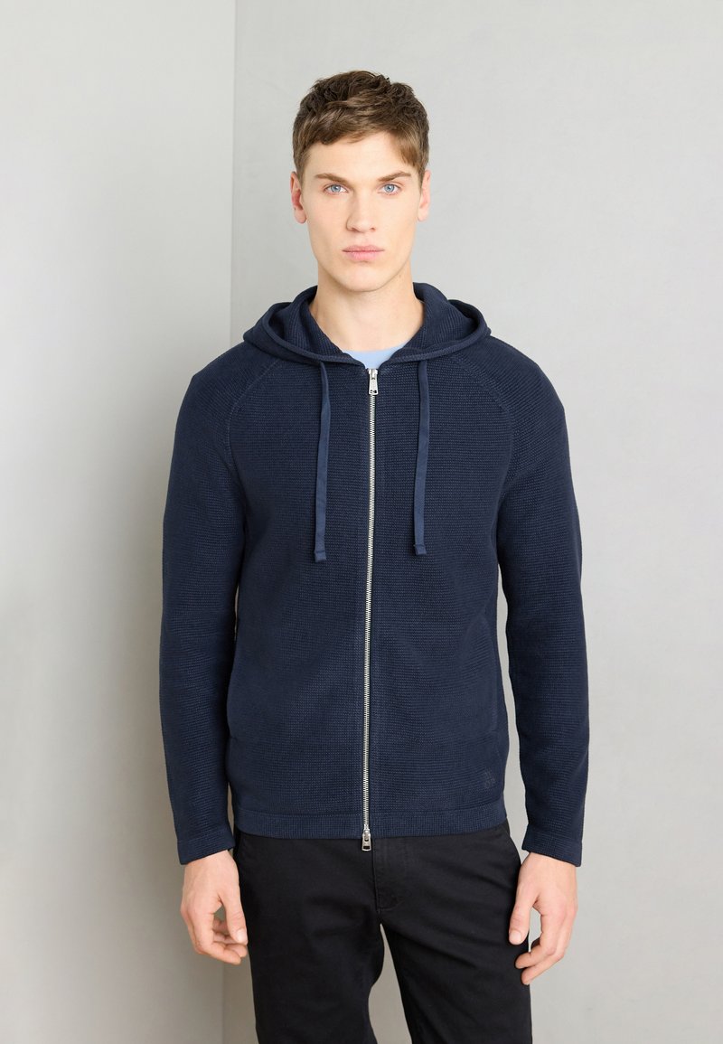 Felpa con zip di colore navy in un tessuto a maglia testurizzato, maniche raglan, cappuccio con cordino e zip liscia; indossata sopra una maglietta azzurra.