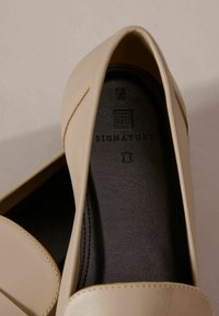 Scarpe slip-on beige con parte superiore in pelle liscia, punta arrotondata e soletta nera contrassegnata con il logo del marchio e la taglia.