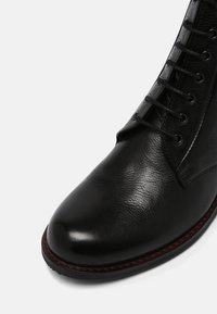 Botín de cuero negro hasta el tobillo con una textura suave, puntera redondeada y costuras rojas en contraste en la suela, con diseño de cordones.