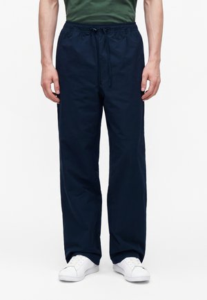 Hombre con pantalones azul marino oscuro con cordón, camisa verde oliva y zapatillas blancas, de pie frente a un fondo liso.
