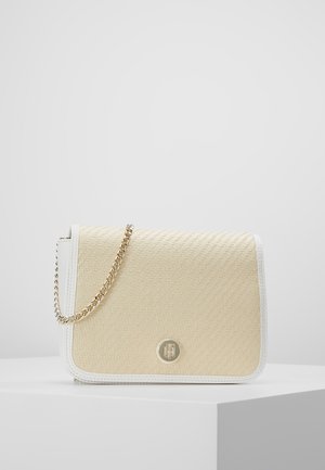 Beżowa pleciona torebka na ramię z białą skórzaną lamówką i złotym łańcuszkowym paskiem, umieszczona na białym piedestale na tle jasnego, jednolitego tła.