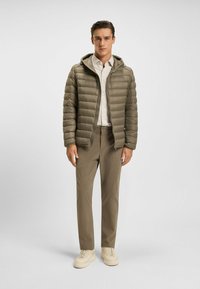 Polstret olivengrøn jakke med hætte, lynlås foran og horisontal quiltning, parret med beige bukser og lyse sneakers.