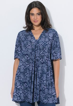 Kvinde med skulderlangt mørkt hår iført en løs blå tunika med paisley-mønster og korte ærmer.