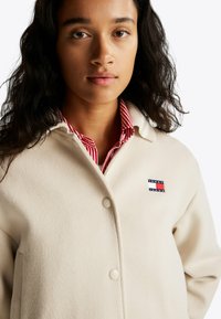 Cremefarvet uldblandingsjakke med knaplukning, der har et lille multifarvet brandlogo på øverste venstre side og et struktureret stof.