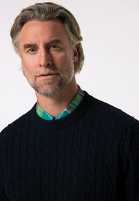 Granatowy sweter w splotie warkoczowym z żeberkowym okrągłym dekoltem, nałożony na koszulę w kolorową kratę z odcieniami zieleni, niebieskiego i fioletu.