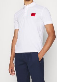 Polo blanc en coton, avec un patch rectangulaire rouge portant le texte "HUGO". Manches courtes, col à revers et coupe ajustée.