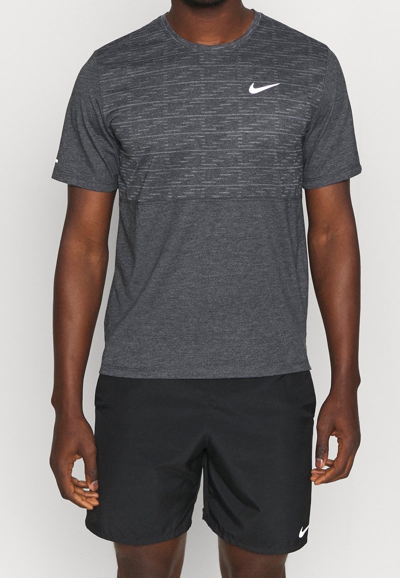 Homme portant un t-shirt Nike gris foncé et un short Nike noir, debout devant un fond uni.