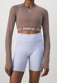 Eine Person trägt ein braunes, langärmliges, cropped Nike-Oberteil und hochgeschnittene, helllila Nike-Radshorts vor einem schlichten Hintergrund.