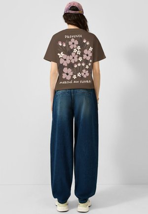 Femme portant un t-shirt marron avec un imprimé floral rose et le texte "Provence Marché Aux Fleurs", jean bleu baggy, chaussures blanches et casquette rose, de dos.
