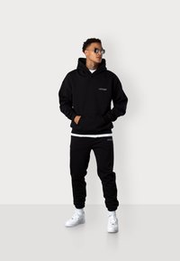 Schwarzes Hoodie und Jogginghose Outfit, mit einer großen Kapuze, Fronttasche und weißen Logodetails. Kombiniert mit weißen Sneakers.