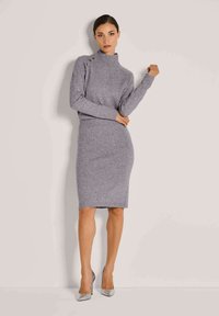 Frau in einem grauen Strickpullover mit Knöpfen an der Schulter und passendem Bleistiftrock, kombiniert mit silbernen, metallischen High Heels.