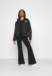 Giacca nera impermeabile con zip, dotata di cappuccio e logo sul petto, abbinata a pantaloni neri a zampa d'elefante e sneakers bianche.