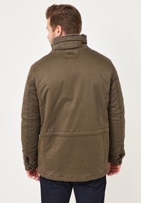 Olivgrüne Jacke aus strukturiertem Stoff mit hohem Kragen und geknöpften Manschetten. Details umfassen ein Logopatch und strukturierte Nähte.