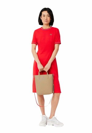 Robe rouge à manches courtes avec un col contrastant ; le modèle tient un sac fourre-tout beige avec de courtes poignées ; des baskets blanches complètent la tenue.