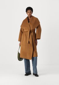 Abrigo de mezcla de lana marrón con un cuello ancho y cinturón, combinado con jeans de mezclilla azules y zapatos negros, sosteniendo un bolso verde.