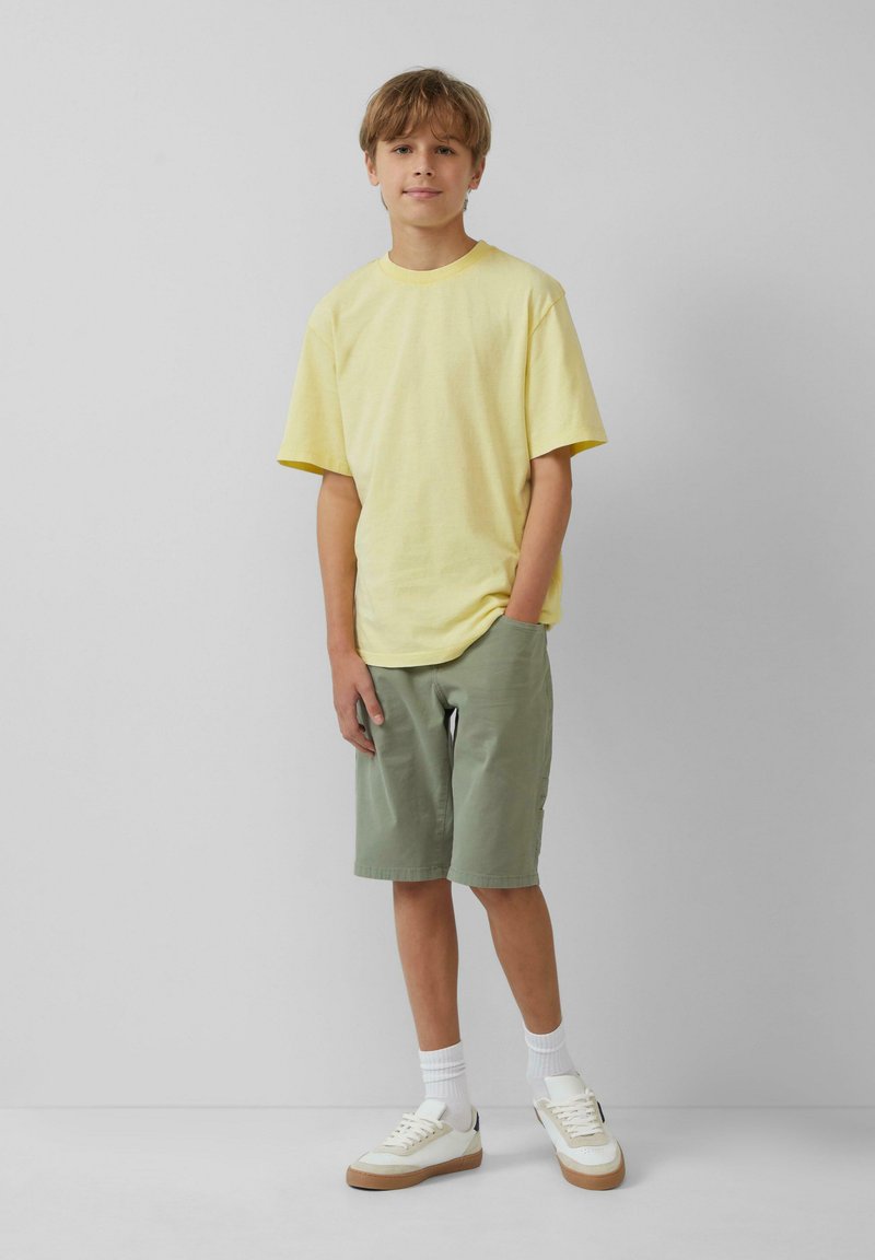 Geel katoenen T-shirt, losse pasvorm, gecombineerd met groene shorts. Witte sneakers met beige accenten en witte crew sokken maken de outfit compleet.
