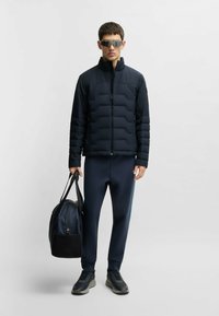 Marinblå puffjacka med kviltat mönster, matchande marinblå träningsoverall och mörka sneakers. Modellen håller en svart duffelväska och bär reflexglasögon.