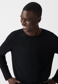 Czarny sweter z długim rękawem i okrągłym dekoltem, wykonany z miękkiego materiału. Modelka stoi z rękami na biodrach, uśmiechając się, na jednolitym białym tle.