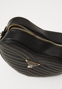 Rund svart lær crossbody-veske med quiltet mønster, gullglidelås og metalllogo-accent foran.