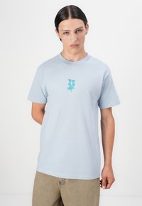 Lyseblå bomulls-T-skjorte med et sentralt akvagrønt grafisk logo. Korte ermer, rund hals, myk tekstur og avslappet passform.