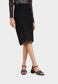 Gonna a tubino in pizzo nero con orlo scallop, abbinata a sandali neri open-toe con cinturini decorati e tacco a blocco.