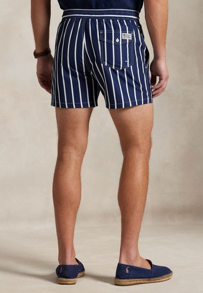 Calções de banho às riscas azul-marinho e branco com bolso traseiro e cintura elástica, combinados com espadrilhas azul-marinho sobre um fundo neutro.