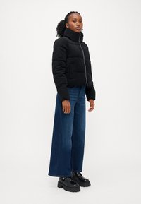 Svart pufferjakke med høy krage og glidelås, parret med vide blå jeans og kraftige sorte sko. Glatt tekstur, avslappet design.