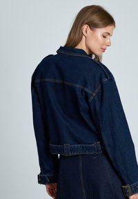Chaqueta de mezclilla azul oscuro con un diseño corto, ajuste holgado y puños abotonados. Las costuras visibles y los detalles de los bolsillos mejoran su estructura.