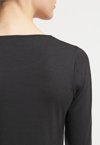 Osoba v jednoduchém černém tričku s dlouhým rukávem, zobrazená zezadu a s mírně otočenou hlavou proti jednoduchému světlému pozadí.