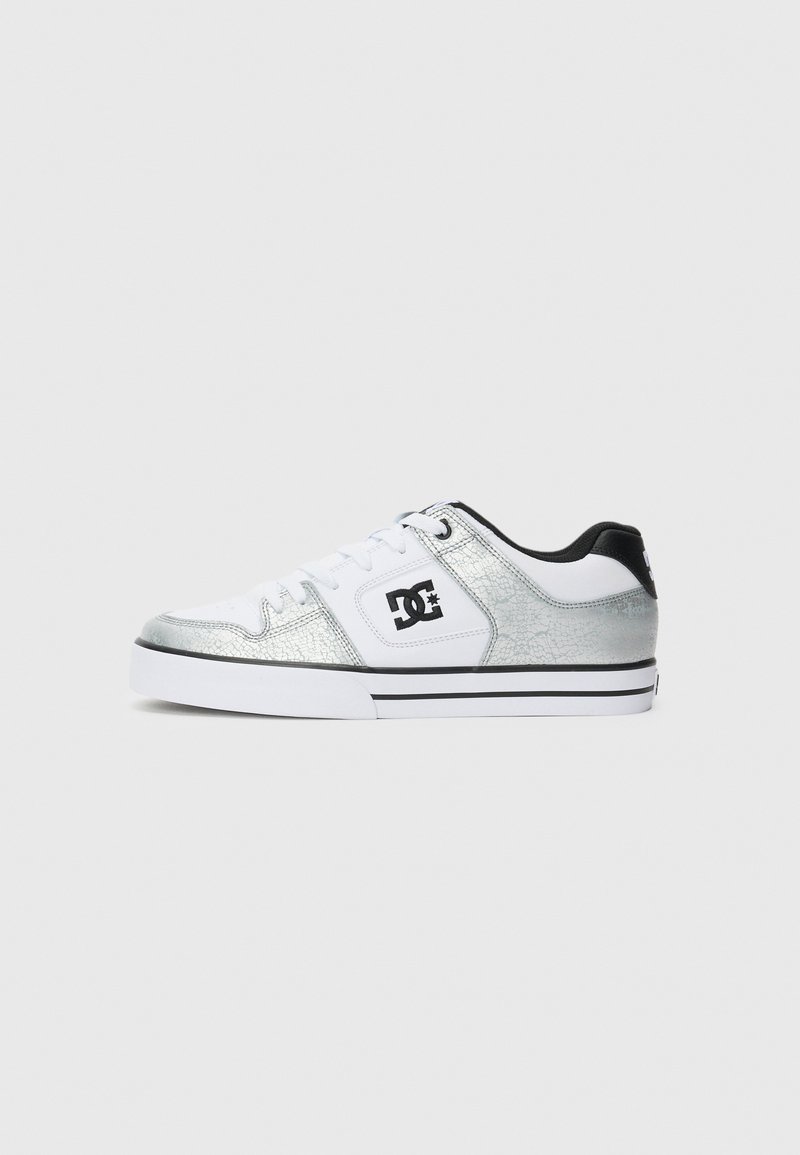 Zapatilla blanca de corte bajo con overlays texturizados en plata, logo negro de DC en el lateral, tira negra en el talón y suela blanca con franja negra.