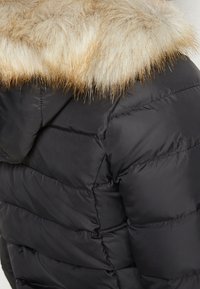 Chaqueta acolchada negra con un diseño de cuadrícula, que presenta un borde de piel sintética removible en la capucha de color marrón claro. Textura suave y brillante.
