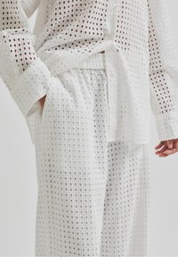 Biały strój z kwadratowym wzorem w oczka, składający się z długiego rękawa koszuli z guzikami oraz dopasowanych szerokich spodni z elastycznym paskiem.