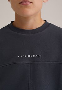 Detailný záber osoby v tmavej mikine s kapucňou, na ktorej je na hrudi bielo vytlačený nápis "BLUE RIDGE DENIM".