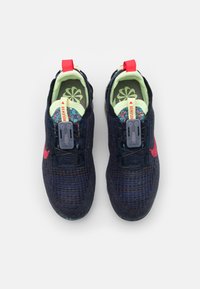 Meripihkan urat olahraga rajutan navy dengan permukaan bertekstur, sol dalam neon hijau, aksen merah, dan mekanisme kait di bagian atas untuk memudahkan pemakaian.