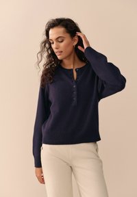 Nainen, jolla on kihara hius, pukeutuneena tummansiniseen nappisveteeseen ja vaaleanbeigeihin housuihin, koskettaa hiuksiaan yksinkertaisen beige-taustan edessä.