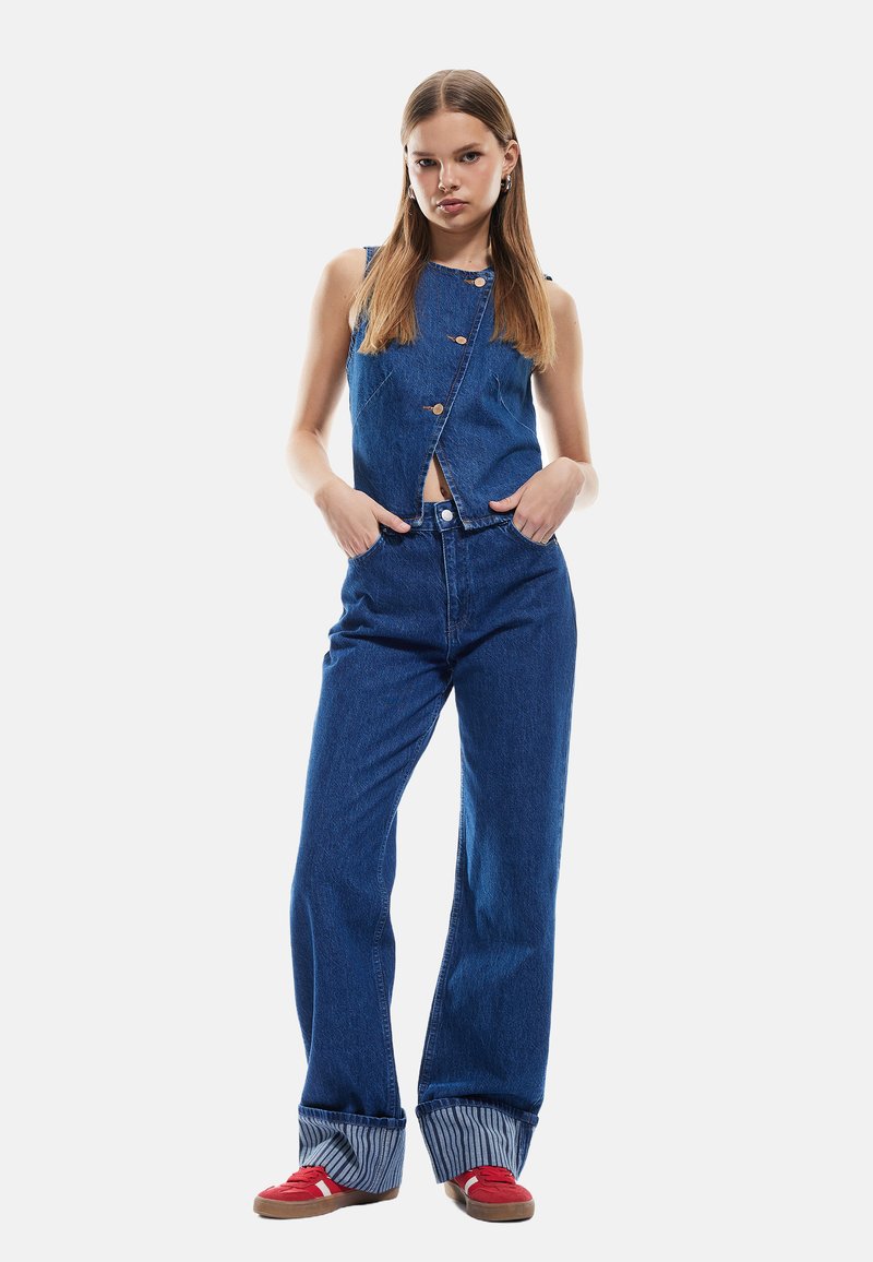Combinaison en denim avec un haut sans manches, des boutons sur le devant et des jambes évasées. Avec des poignets rayés et portée avec des baskets rouges.