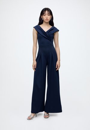 Kvinde iført en marineblå jumpsuit med vide ben og off-shoulder V-hals samt sølvfarvede hælede sandaler, stående mod en ensfarvet lys baggrund.