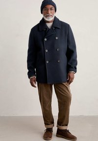 Cappotto di lana doppiopetto blu navy con bottoni grigi, indossato sopra un maglione chiaro. Abbinato a pantaloni in velluto a coste marroni e stivali stringati marroni.