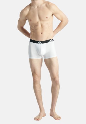 Oberkörperfreier Mann in weißen Boxershorts mit schwarzem Bund und sportlichem Logo, der barfuß vor einem schlichten Hintergrund steht.