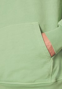 Sudadera verde hecha de tela suave, con un bolsillo frontal y puños acanalados. Detalle sutil de marca en la tela.