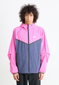 Ung person med krøllet hår iført en lyserød og grå Nike zip-up jakke med hætte og sorte bukser, der står foran en hvid baggrund.