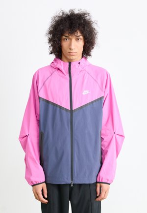 Young person with curly hair wearing a pink and gray Nike zip-up jacket with hood and black pants, standing against a white background.