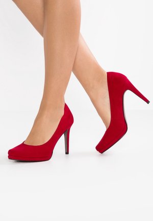 Czerwone zamszowe buty na wysokim obcasie z okrągłym noskiem i platformą. Obcasy są wąskie i czarne, co podkreśla intensywny kolor.