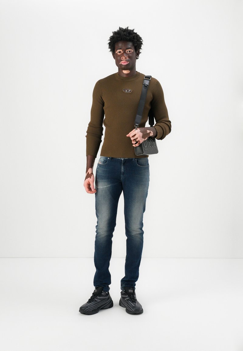 Brauner gerippter Pullover mit Logo, blaue Slim-Fit-Jeans und schwarze Sneakers. Das Modell trägt eine kleine schwarze Schultertasche mit geprägtem Detail.