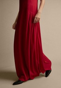 Robe maxi rouge fluide en tissu léger, dotée de panneaux texturés et de plis, assortie à des chaussures noires à bout pointu.