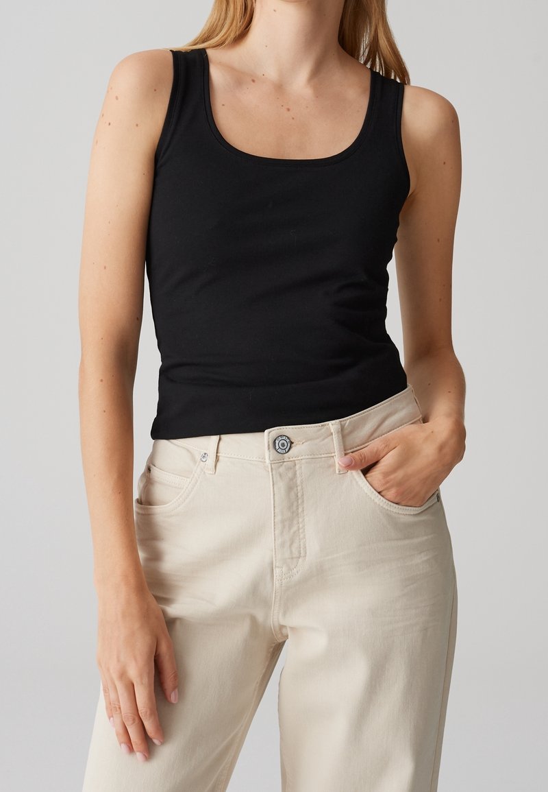 Femme portant un haut noir sans manches et un pantalon taille haute couleur crème, une main dans la poche et fond neutre.