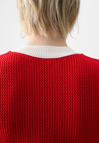Personne aux cheveux blonds courts, vue de dos, portant un pull en tricot rouge avec un col blanc et de grosses boucles d'oreilles en forme d’anneaux sur un fond blanc.